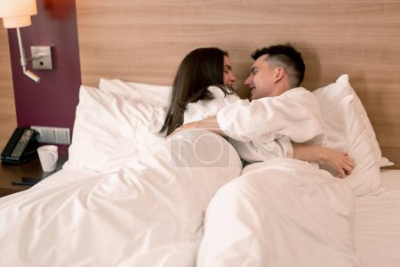 Téléchargez les photos : Portrait jeune couple en lune de miel dans la chambre d'hôtel couché sur le lit peignoirs blancs embrasser heureux amoureux concept de voyage - en image libre de droit