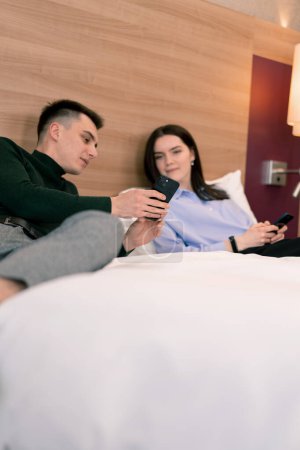 Photo for Young couple in the room of a luxury hotel room, lovers lying on the bed watching funny videos on phone travel concept - Royalty Free Image