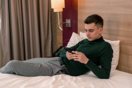 Foto de Un joven en la habitación de una habitación de hotel de lujo tiene un teléfono inteligente en sus manos y resuelve problemas de negocios después del viaje - Imagen libre de derechos