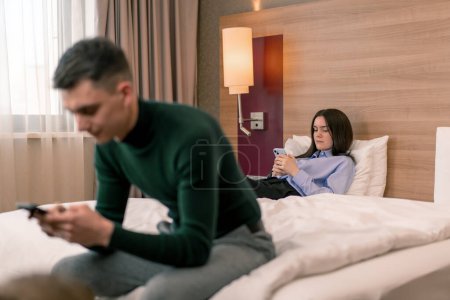 Téléchargez les photos : Jeune couple dans la chambre d'une chambre d'hôtel de luxe, les gens tiennent smartphones dans leurs mains ne communiquent pas entre eux Voyage - en image libre de droit