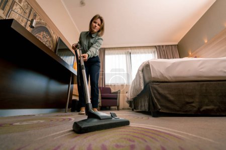 Foto de Muchacha joven trabajadora de limpieza en hotel chica limpieza habitación aspiradora alfombra en habitación de lujo concepto limpieza y hospitalidad - Imagen libre de derechos