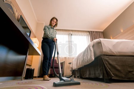 Photo for Young girl housekeeping worker in hotel girl cleaning room vacuuming carpet in luxury room concept cleanliness and hospitality - Royalty Free Image