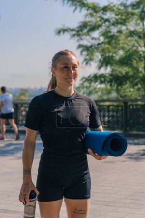Foto de Retrato de una joven deportista sonriente sosteniendo una campana de agua y una botella de agua en sus manos antes de comenzar un entrenamiento al aire libre en el parque - Imagen libre de derechos