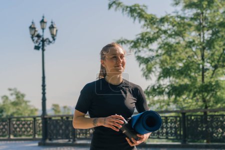 Téléchargez les photos : Portrait d'une jeune sportive souriante tenant une kettlebell et une bouteille d'eau dans ses mains avant de commencer une séance d'entraînement en plein air dans le parc - en image libre de droit