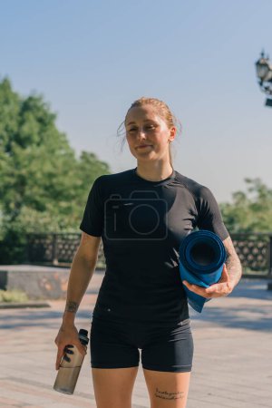 Foto de Retrato de una joven deportista sonriente sosteniendo una campana de agua y una botella de agua en sus manos antes de comenzar un entrenamiento al aire libre en el parque - Imagen libre de derechos