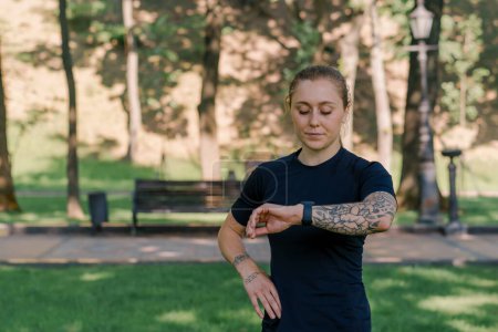 Téléchargez les photos : Portrait d'une jeune sportive vérifiant le temps et le pouls d'une montre intelligente après une séance d'entraînement dans le parc concept de mode de vie sain - en image libre de droit