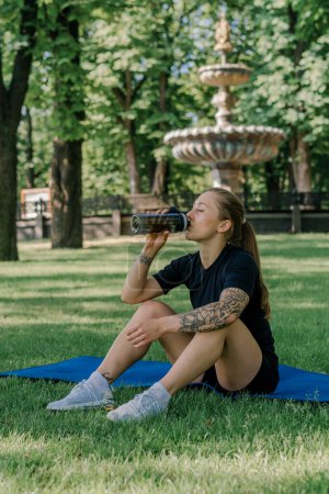 Photo for Portrait of young satisfied sports girl resting on sports mat drinking water outdoors after training park healthy lifestyle concept - Royalty Free Image