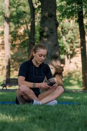 Téléchargez les photos : Portrait d'une jeune athlète sur un tapis de sport se reposant après l'entraînement dans le parc avec un smartphone dans ses mains le concept de mode de vie sain - en image libre de droit