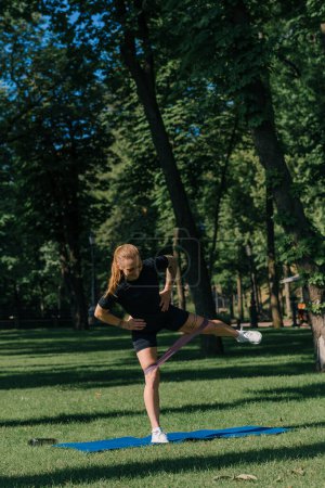 Téléchargez les photos : Jeune fille sportive faisant des exercices physiques sur un tapis de sport entraînement avec des élastiques sur ses jambes à l'extérieur dans le parc concept de mode de vie sain - en image libre de droit