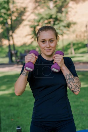 Photo for Portrait of a young satisfied sports girl doing exercises with dumbbells outdoors during a workout in park healthy lifestyle concept - Royalty Free Image