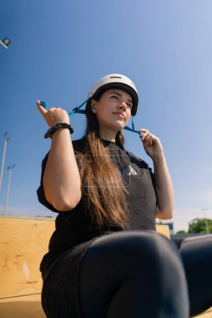 Photo for Portrait of young concentrated hipster girl against sky background in skate park putting safety helmet before starting roller skating - Royalty Free Image