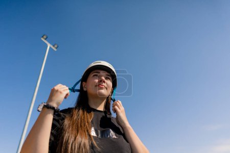 Téléchargez les photos : Portrait de jeune fille hipster concentré sur fond de ciel dans le skate park mettant casque de sécurité avant de commencer le patinage à roulettes - en image libre de droit