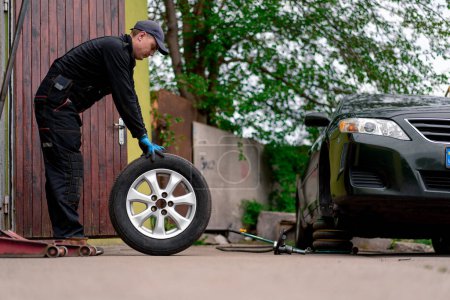 Foto de Mecánico automático en una tapa cambiando una rueda de un coche negro usando un taladro en el taller de neumáticos en la calle detallando las reparaciones del coche - Imagen libre de derechos
