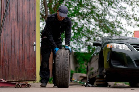 Téléchargez les photos : Mécanicien automobile dans un capuchon changer une roue d'une voiture noire à l'aide d'une perceuse dans l'atelier de pneus sur la rue détaillant les réparations de voiture - en image libre de droit