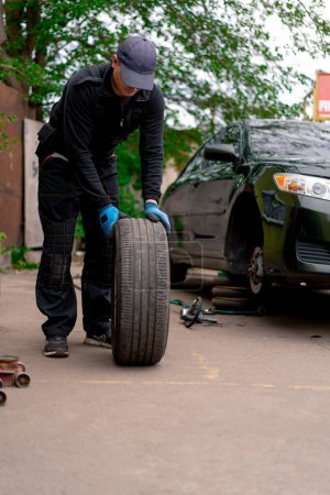 Photo for Auto mechanic in a cap changing a wheel from a black car using a drill in tire shop on the street detailing car repairs - Royalty Free Image