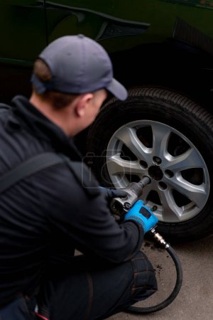 Photo for Auto mechanic in a cap changing a wheel from a black car using a drill in tire shop on the street detailing car repairs - Royalty Free Image