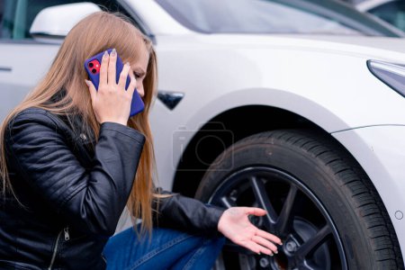 Foto de Molesto chica atractiva agachada junto a la rueda de un coche en el lado de la carretera hablando por teléfono cerca de coche roto y pidiendo ayuda - Imagen libre de derechos