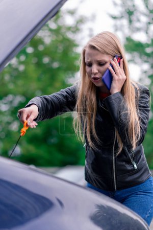 Téléchargez les photos : Fille attrayant bouleversé se tient à côté de la voiture avec un capot ouvert sur le côté de la route parler au téléphone vérifier le niveau d'huile et appeler à l'aide - en image libre de droit