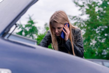 Photo for Upset attractive girl stands next to a car with an open hood on the side of the road talking on the phone, standing by broken car and calling for help - Royalty Free Image