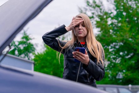 Foto de Una chica molesta con el teléfono en las manos está cerca de la capucha abierta de su coche azul de lujo y trata de entender lo que se rompe en ella - Imagen libre de derechos