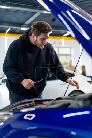 Photo for A man with wrench in his hands stands at the open hood of his luxurious blue car that has broken down and thinks how to fix it - Royalty Free Image