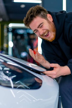 Téléchargez les photos : Un homme souffre en train de se pincer les doigts sous le capot de sa voiture de luxe blanche dans un parking ou un service de voiture en gros plan - en image libre de droit
