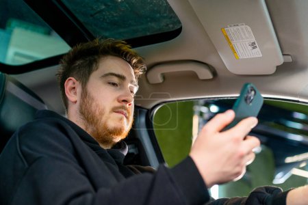 Foto de Un hombre se sienta al volante de su coche de lujo con un teléfono en sus manos y lo mira en un estacionamiento o en el servicio de automóviles - Imagen libre de derechos