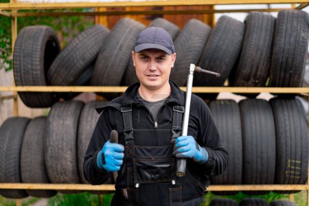 Photo for Portrait of a serious man Auto mechanic showing Thumb signal with a cylinder wrench in his hand on the background of car tires detailing auto repair - Royalty Free Image