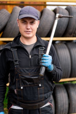Photo for Portrait of a serious man Car mechanic with balloon wrench in his hand dressed in work uniform on the background of car tires detailing car repair - Royalty Free Image