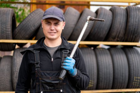 Foto de Retrato de un hombre serio Mecánico de coche con llave de globo en la mano vestido con uniforme de trabajo en el fondo de los neumáticos de coche detallando la reparación del coche - Imagen libre de derechos