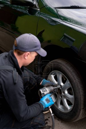 Téléchargez les photos : Mécanicien automobile dans un capuchon changer une roue d'une voiture noire à l'aide d'une perceuse dans l'atelier de pneus sur la rue détaillant les réparations de voiture - en image libre de droit