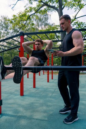 Foto de Un entrenador de fitness con una tableta en sus manos entrena a un joven atleta bombeando sus abdominales en la calle en el campo de deportes apoyo de motivación - Imagen libre de derechos