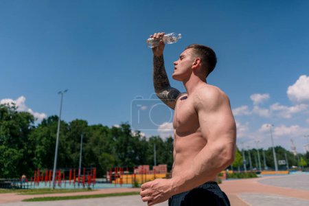 Téléchargez les photos : Jeune bodybuilder sportif debout sur le terrain de sport obtenir éclaboussé de chaleur de l'eau après la formation de rue belle sexy pompé corps - en image libre de droit