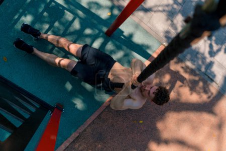 Téléchargez les photos : Jeune sportif avec un torse nu tire vers le haut sur une corde pendant l'entraînement en plein air sur le terrain de sport sexy pompé corps - en image libre de droit