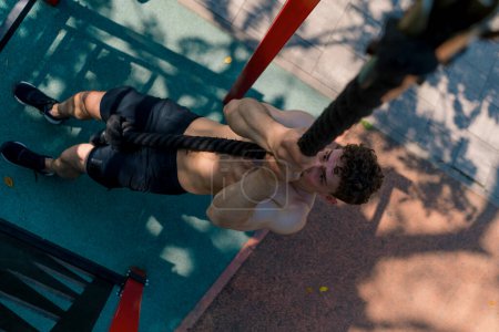 Téléchargez les photos : Jeune sportif avec un torse nu tire vers le haut sur une corde pendant l'entraînement en plein air sur le terrain de sport sexy pompé corps - en image libre de droit