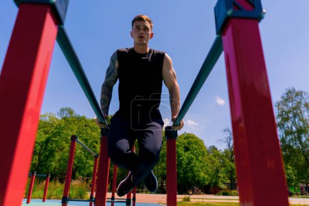 Foto de Retrato de un joven deportista haciendo ejercicio en barras paralelas balanceando sus brazos y hombros haciendo flexiones campo de deportes durante el entrenamiento callejero - Imagen libre de derechos