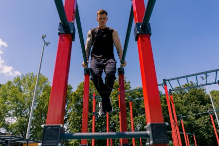Téléchargez les photos : Portrait d'un jeune sportif travaillant sur des barres parallèles balançant ses bras et ses épaules faisant des pompes terrain de sport pendant l'entraînement de rue - en image libre de droit