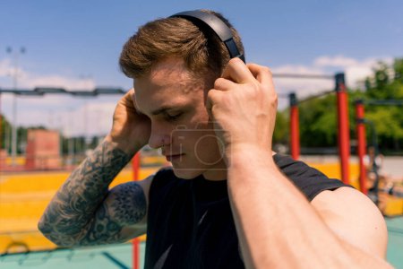 Photo for Portrait of a young sweaty sportsman with tattoos and piercings wearing headphones on the sports ground outdoor training in park motivation - Royalty Free Image