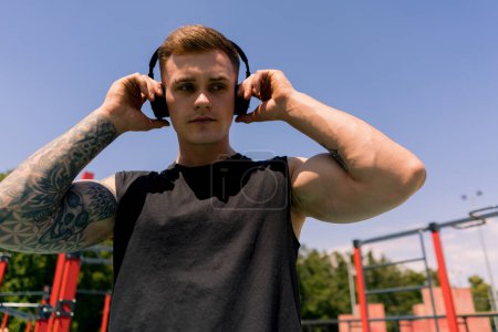 Photo for Portrait of a young sweaty sportsman with tattoos and piercings wearing headphones on the sports ground outdoor training in park motivation - Royalty Free Image