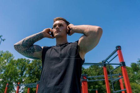 Téléchargez les photos : Portrait d'un jeune sportif en sueur avec des tatouages et des piercings portant des écouteurs sur le terrain de sport entraînement en plein air dans la motivation du parc - en image libre de droit