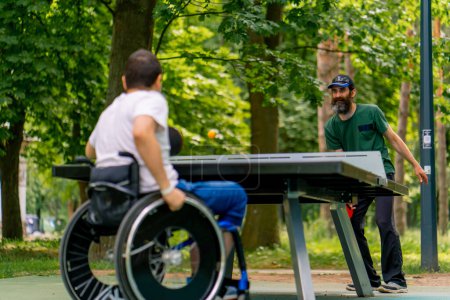 Foto de Inclusividad Un hombre discapacitado en silla de ruedas juega ping pong con un hombre mayor en un parque de la ciudad contra el telón de fondo de los árboles - Imagen libre de derechos