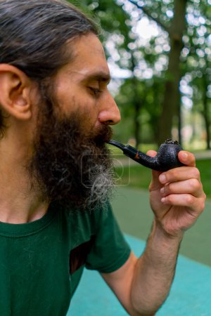 Foto de Retrato de un anciano de pelo largo y barba gris fumando una pipa en el parque de la ciudad sobre el fondo de los árboles - Imagen libre de derechos