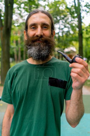 Photo for Portrait of an aged man with long hair and gray beard smoking a pipe in city park against the background of trees - Royalty Free Image