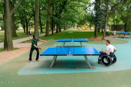 Photo for Inclusiveness A disabled man in a wheelchair plays ping pong against an old man with a gray beard in city park - Royalty Free Image