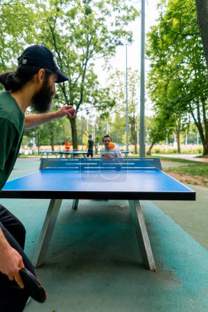 Foto de Inclusividad anciano jugando ping-pong contra un hombre con discapacidad que está en una silla de ruedas en el parque de la ciudad - Imagen libre de derechos