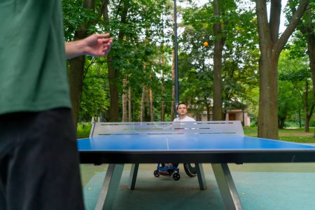 Foto de Inclusividad Un hombre discapacitado en silla de ruedas juega ping pong contra un anciano con barba gris en el parque de la ciudad - Imagen libre de derechos