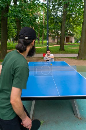 Téléchargez les photos : Inclusivité vieil homme jouant au ping-pong contre un homme handicapé qui est en fauteuil roulant dans un parc municipal - en image libre de droit