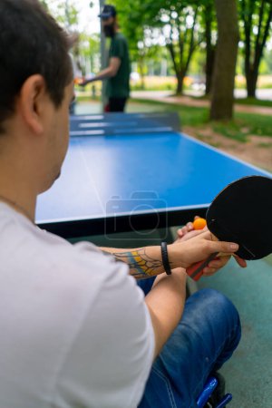 Foto de Inclusividad Un hombre discapacitado en silla de ruedas juega ping pong en un parque de la ciudad contra el telón de fondo de los árboles - Imagen libre de derechos