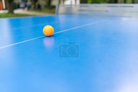 Téléchargez les photos : Une balle de tennis orange repose sur une table de tennis bleue à côté d'un filet dans un parc municipal le concept de jouer au ping-pong à l'extérieur - en image libre de droit