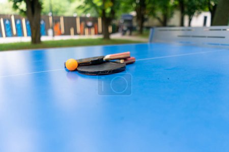 Foto de Dos raquetas de tenis y una pelota de tenis naranja se encuentran en una mesa de tenis azul junto a una red en un juego de ping pong en el parque de la ciudad - Imagen libre de derechos
